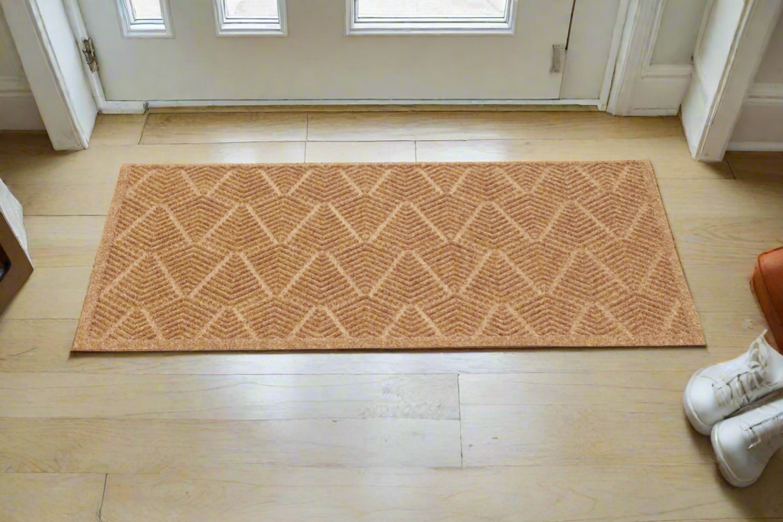 A rectangular, beige recycled PET doormat with a geometric pattern is placed on a wooden floor inside a home, with shoes at the bottom of the image.