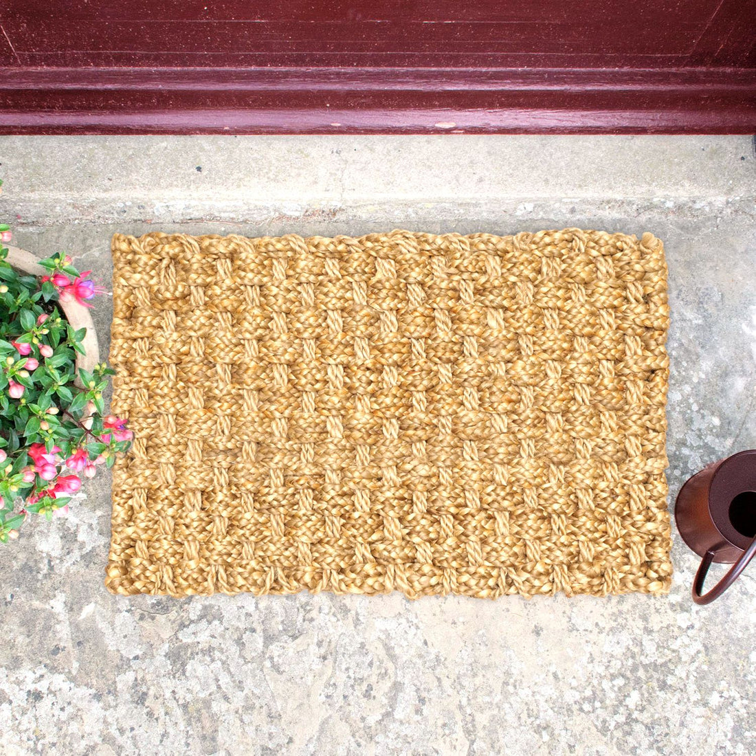 A biege braided jute mat with a traditional geometric pattern, placed on a doorstep with a plant on one side and a brown pot on the other.