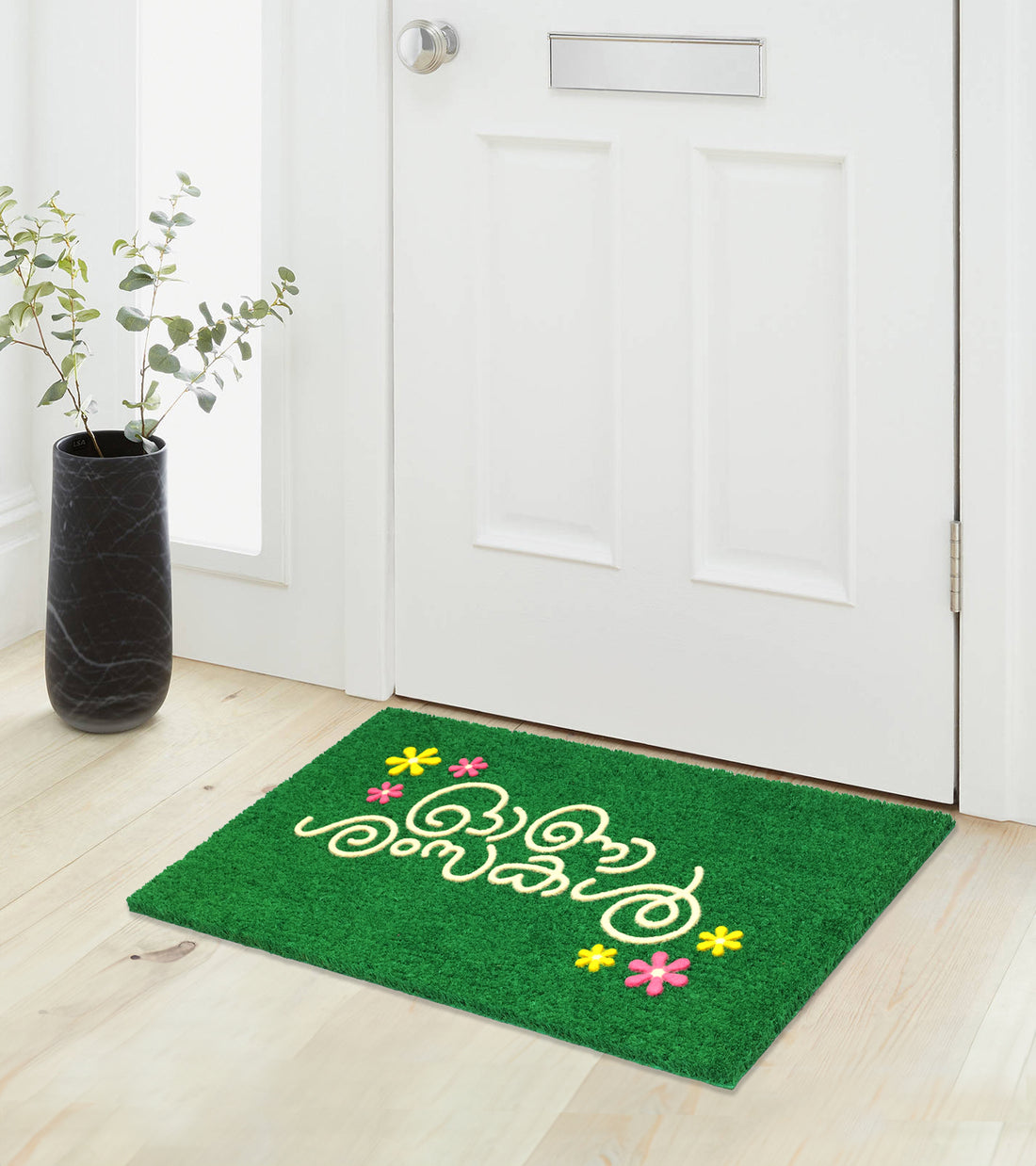 A green doormat with 'Happy Onam' printed in Malayalam and decorative flowers, placed on a doorstep.