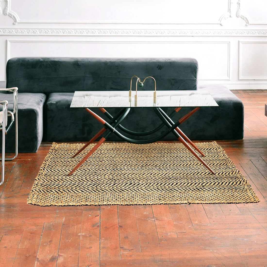 A rectangular jute rug with a chevron pattern in a herringbone weave design, placed on a wooden floor in a living room setting with a black couch, a white and gold coffee table, and a black metal frame chair.