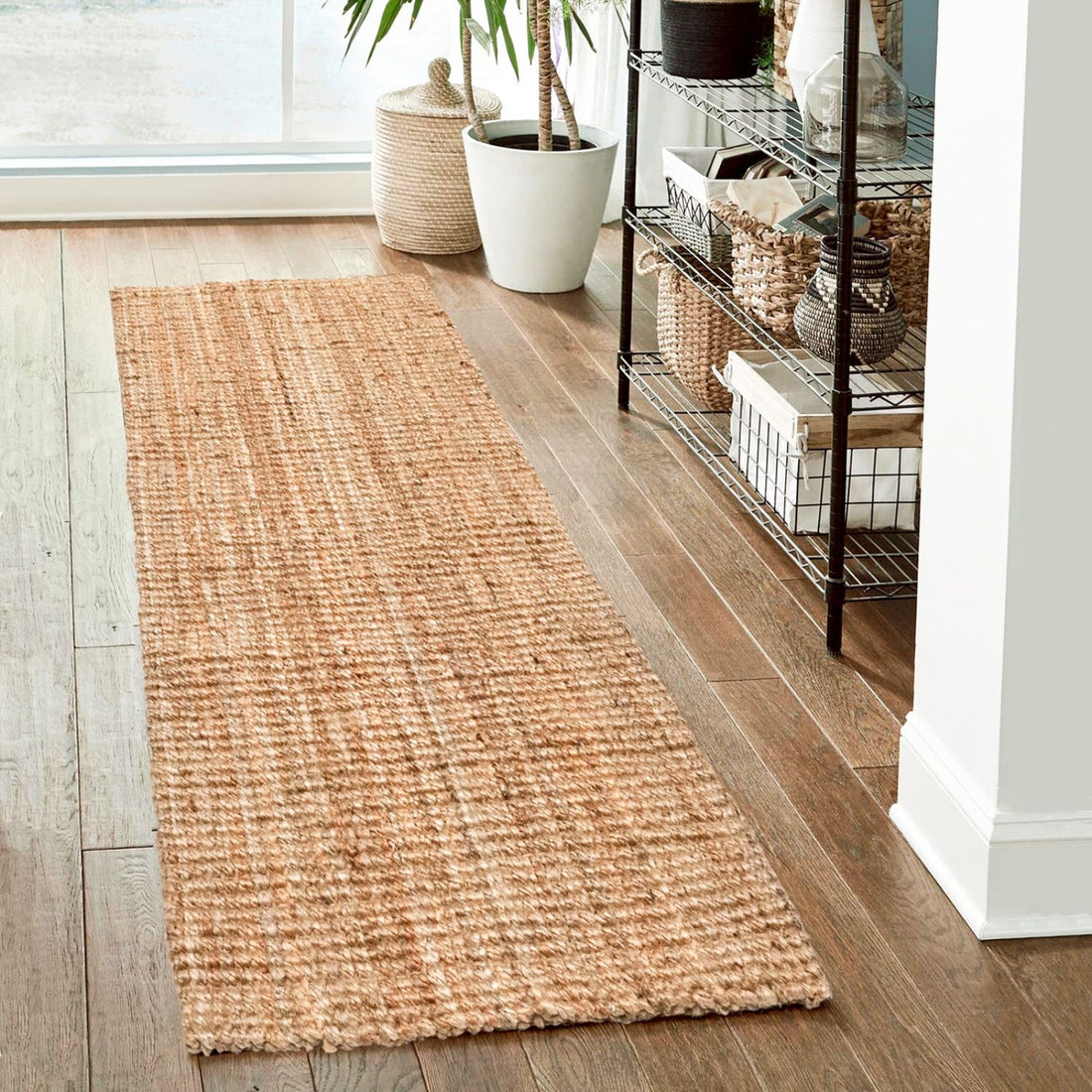Woven runner jute rug on a wooden floor with a plant and storage rack in the background.