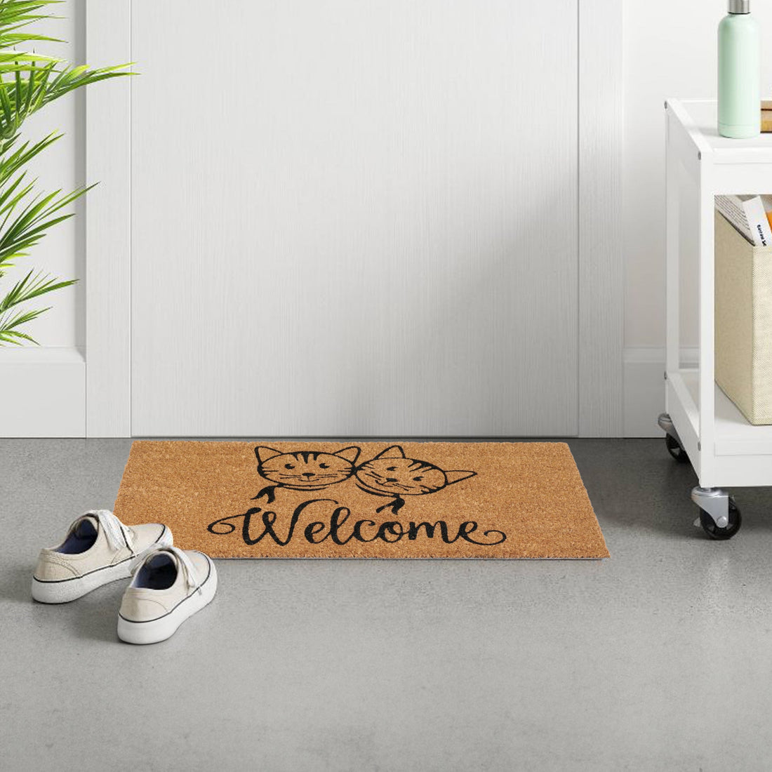 A rectangular natural coir doormat with a printed design featuring two cats and the word 'WELCOME' on a gray floor, next to a white door and a pair of shoes.