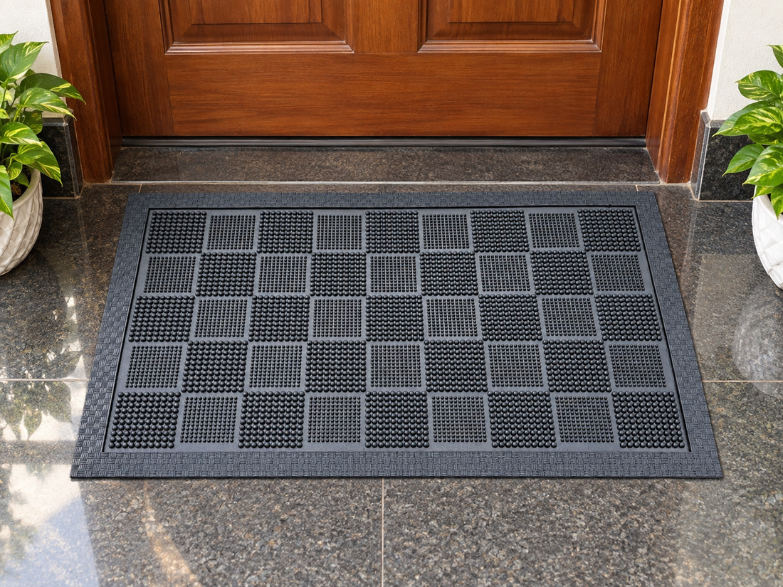 
                      
                        A rectangular black rubber doormat with a textured grid pattern, positioned in front of a wooden door on a polished stone floor, flanked by potted green plants.
                      
                    