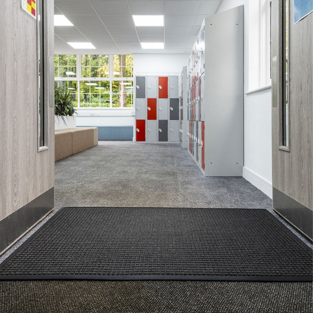 A commercial entryway featuring a black OnlyMat commercial quickdry sanitize mat placed on the floor, with a patterned wall and lockers in the background.