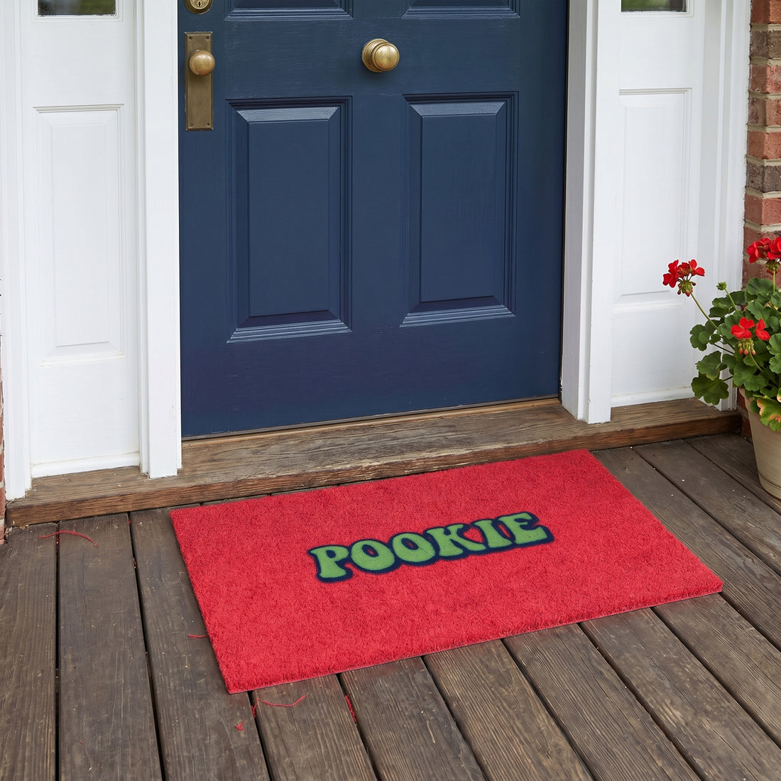 A red rectangular doormat with the word 'POOKIE' in green letters outlined in black, placed on a wooden porch in front of a blue front door.