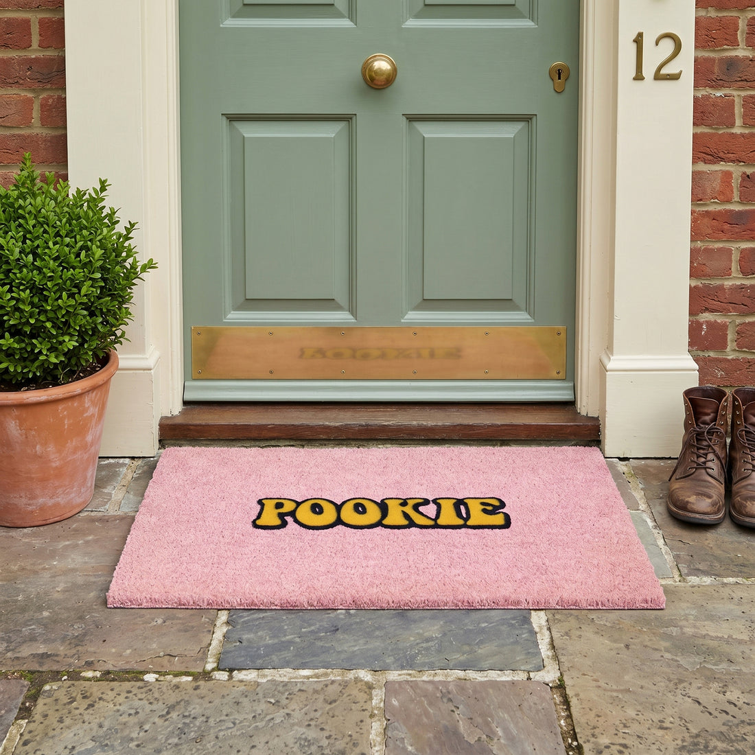 
                      
                        A pink rectangular doormat with the word 'POOKIE' in yellow letters outlined in black, placed in front of a green front door on a stone patio. A potted plant is on the left, a pair of brown boots is on the right, and the house number 12 is visible on the wall.
                      
                    
