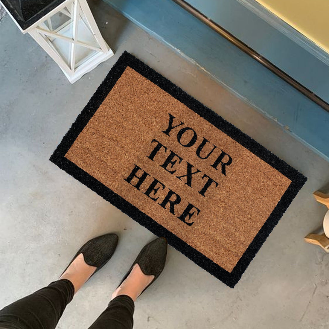 A personalized coir doormat with custom text printed in black. The text 'YOUR TEXT HERE' is displayed on the mat, which is placed on the floor and has a person's feet in black shoes positioned on top of it.