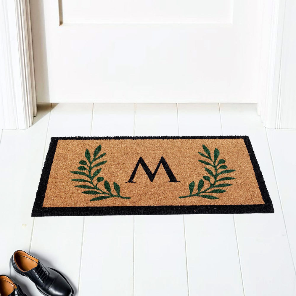 A custom monogram coir doormat with a green leaf border design, placed on a white doorstep, with a pair of shoes to the side.