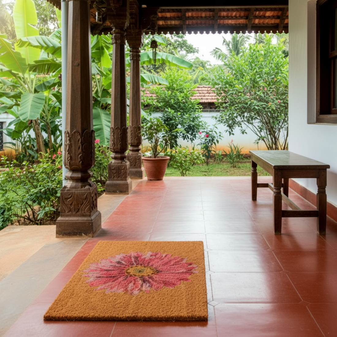 Traditional outdoor veranda with a floral coir doormat, wooden bench, and lush green garden.