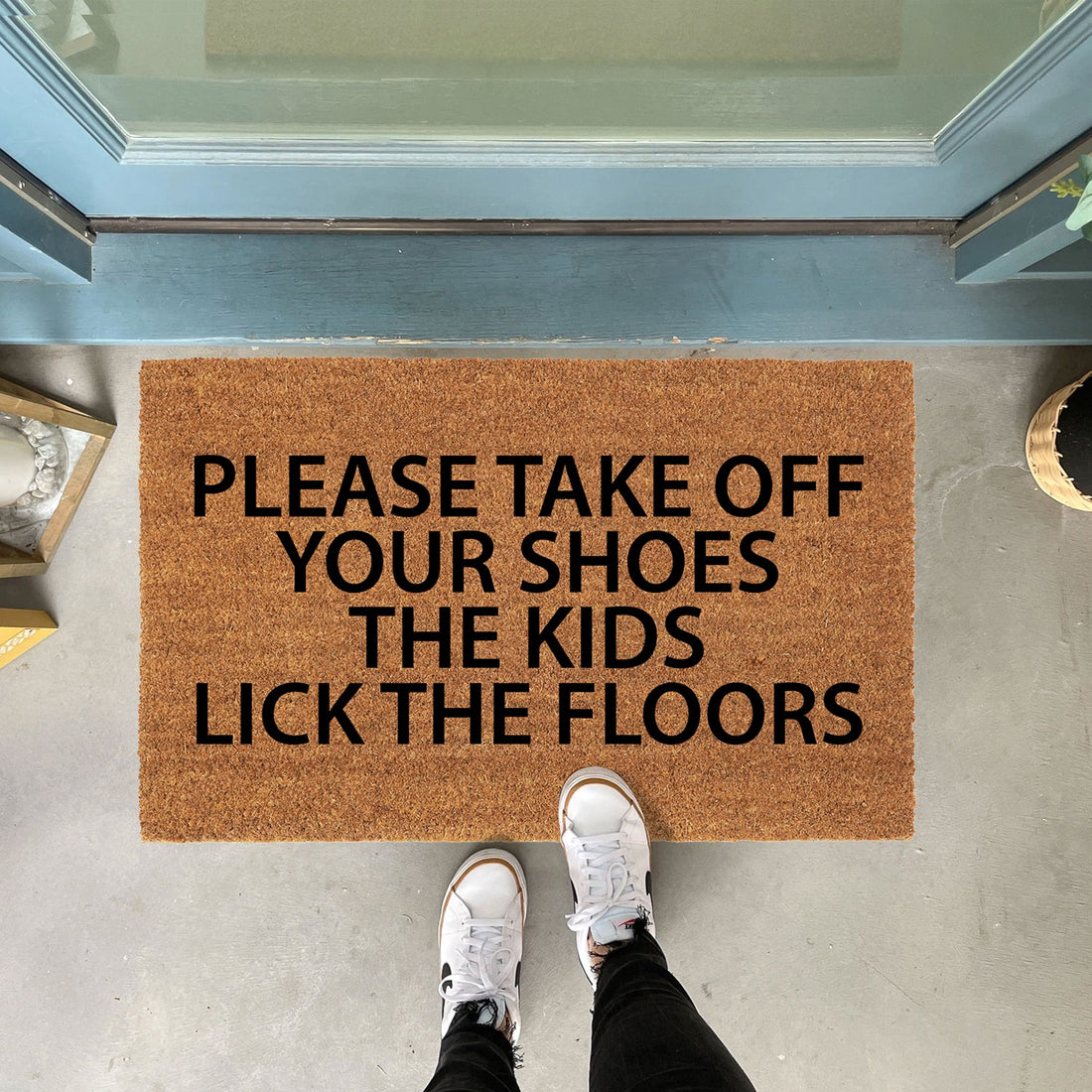 
                      
                        A natural coir doormat with a printed message 'PLEASE TAKE OFF YOUR SHOES THE KIDS LICK THE FLOORS' in bold letters. The doormat is placed at the entrance of a home, and a person's feet wearing white sneakers are shown stepping onto it.
                      
                    