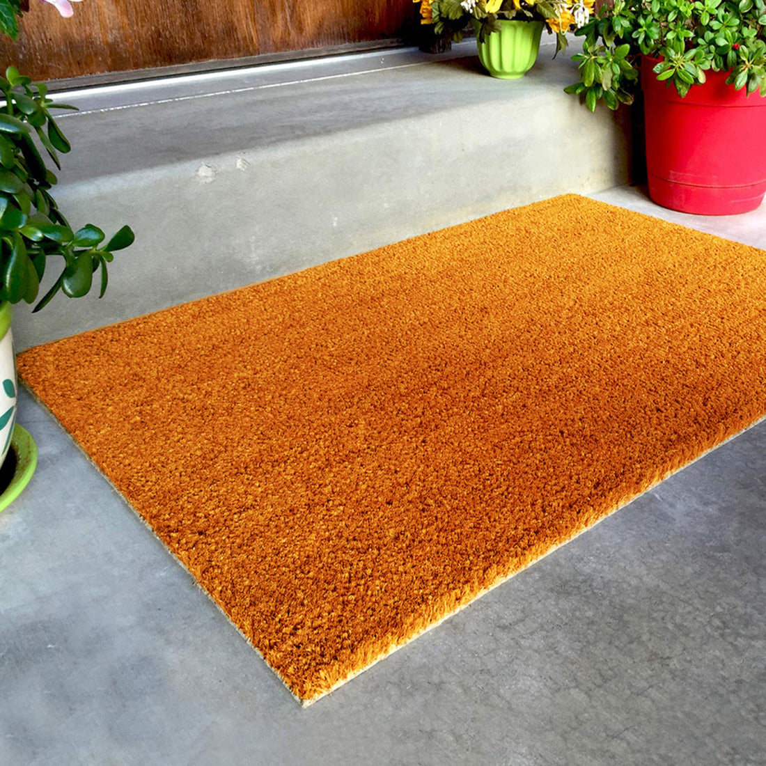 A natural brown coir doormat placed on a concrete step with a potted plant and flowers in the background.