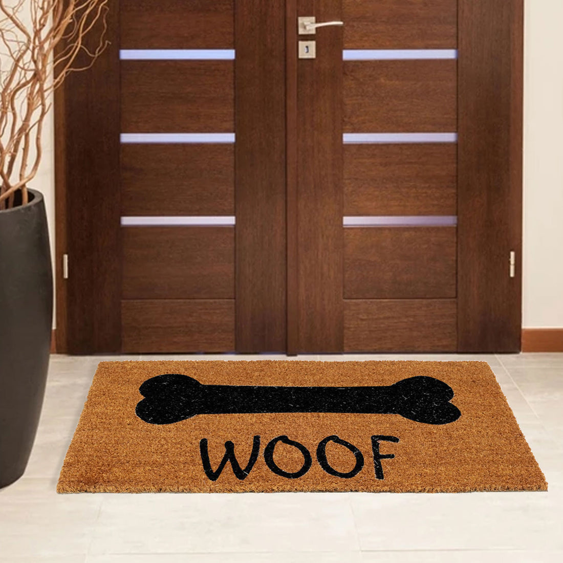 Rectangular coir doormat with a printed design of a bone and the word 'WOOF' on it, placed on the floor in front of a wooden door.