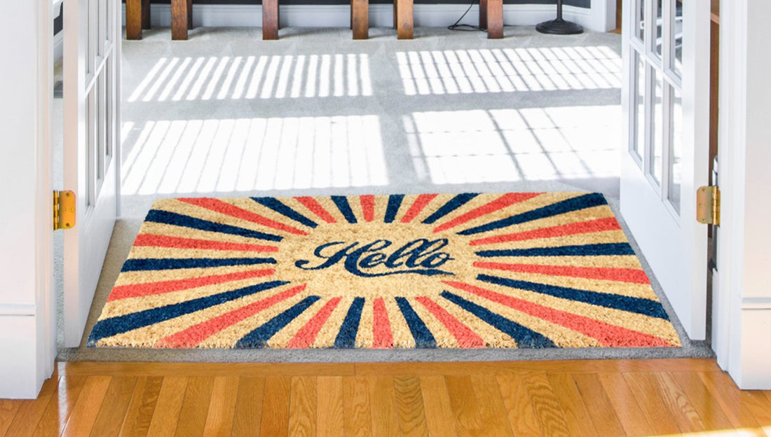 A rectangular natural coir door mat with a printed design that reads 'Hello' in the centre, featuring a colourful sunburst pattern with orange, blue, and red colors.
