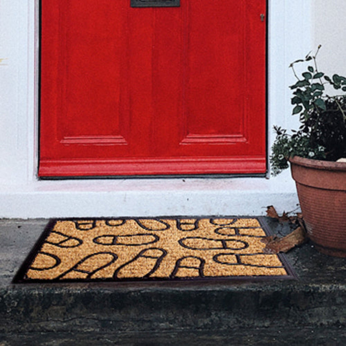 
                      
                        A rectangular natural coir floor mat with a black border and moulded foot mark design, placed on a doorstep with a red door in the background and a potted plant to the side.
                      
                    