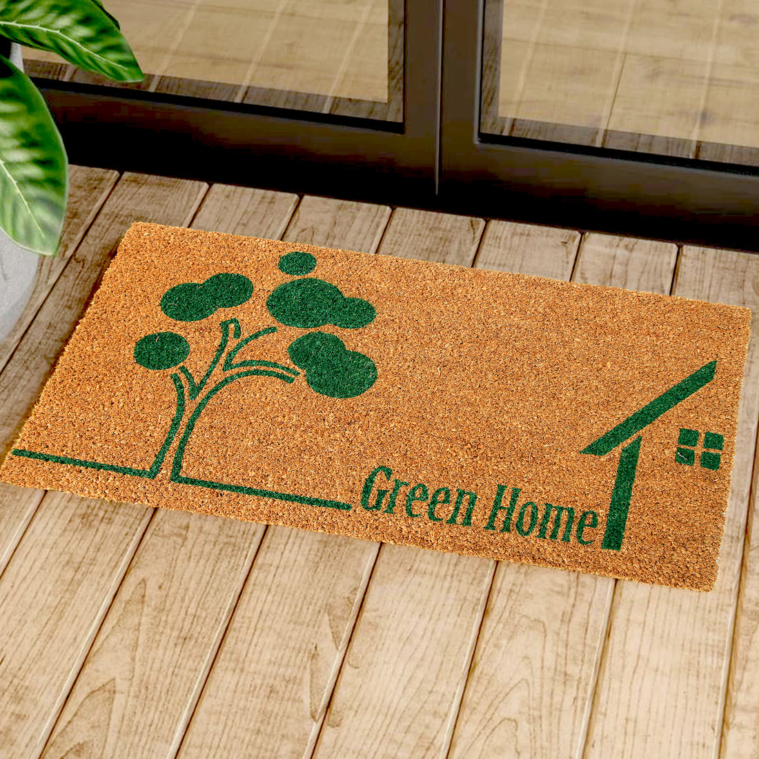 Rectangular coir doormat with a green tree and house design, placed on a wooden floor, with a plant in the background.