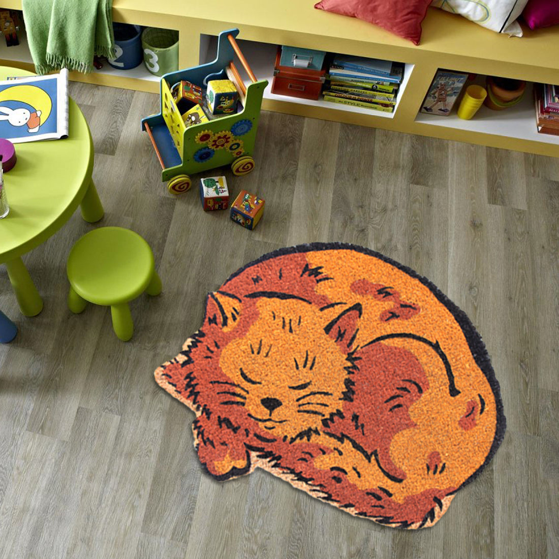 A cat-shaped coir doormat with anti-skid vinyl backing, featuring colourful stencil work, is placed on a floor with children's toys around it.