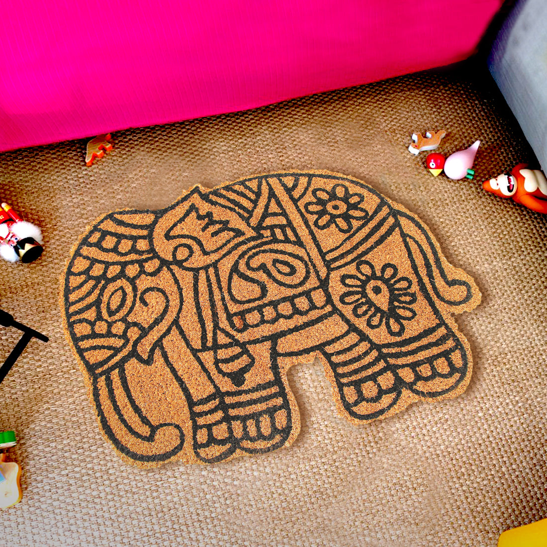 An elephant-shaped coir floor mat with a geometric and floral pattern, placed on a living room floor next to a pink couch with decorative items around it.