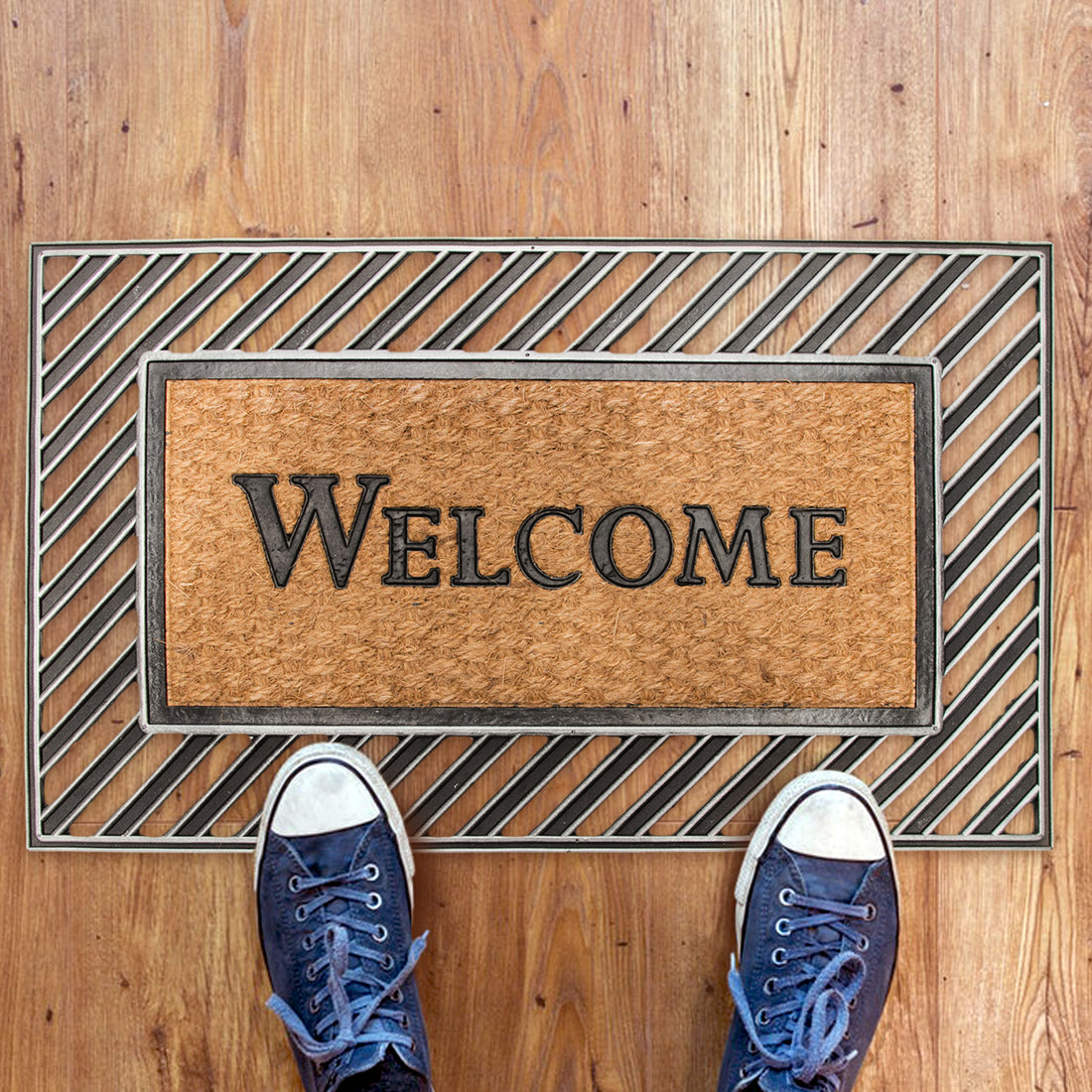 A rectangular coir welcome mat with rubber moulded edges, featuring the word 'WELCOME' in bold capital letters, placed on a wooden floor with a pair of blue sneakers at the bottom of the image.