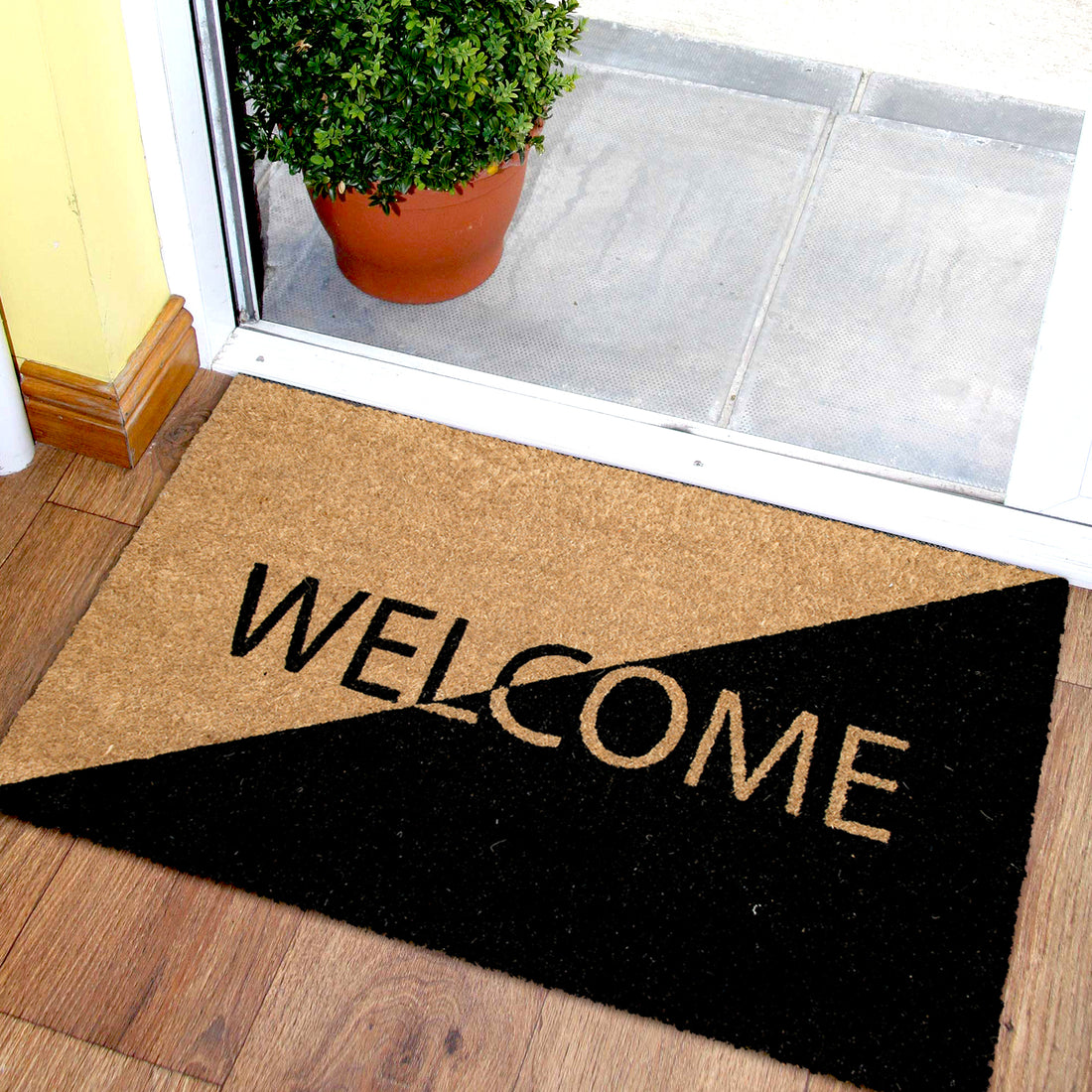 A rectangular black doormat with the word 'Welcome' printed in large black letters, placed on the floor in front of a door, with a potted plant to the left and a wooden floor visible to the side.