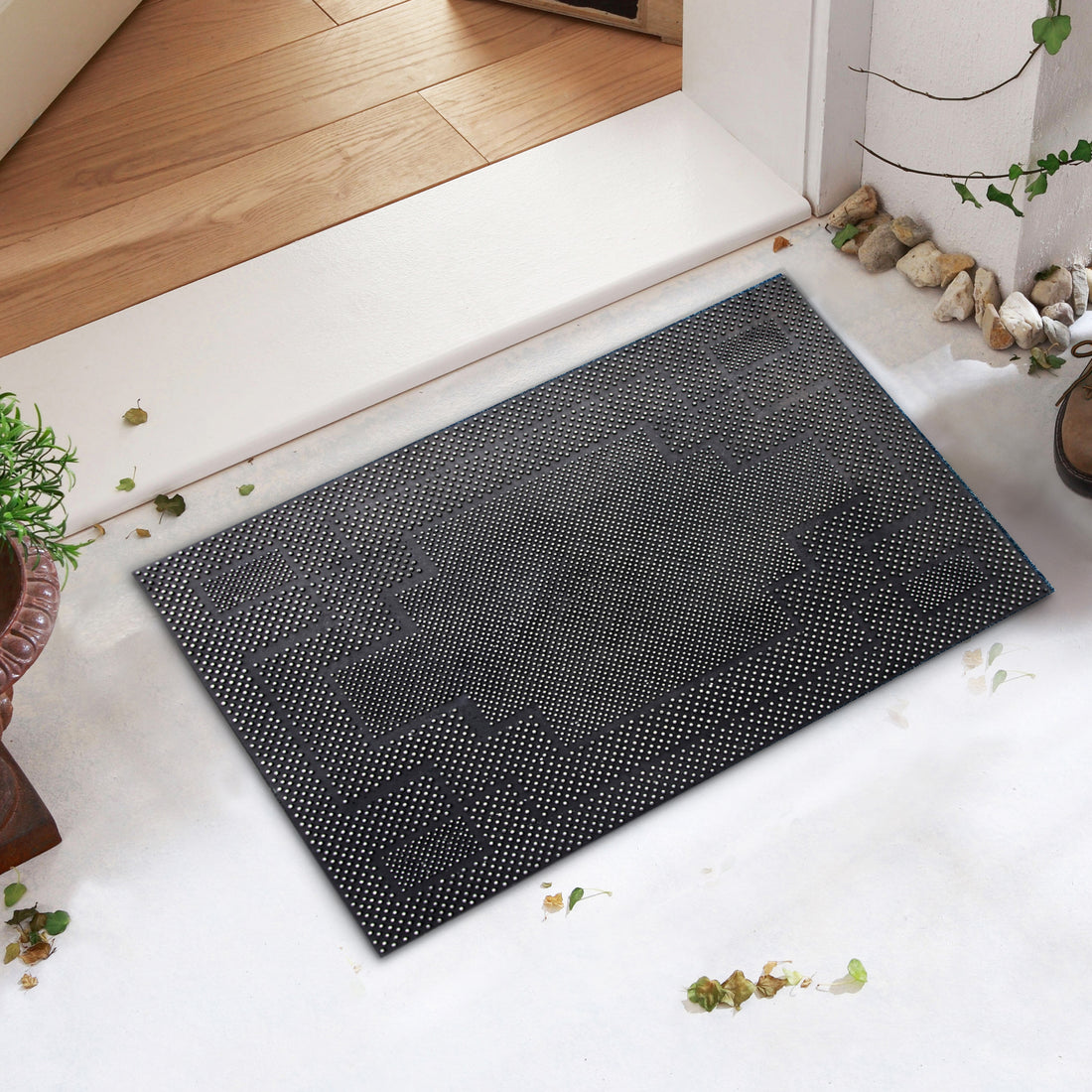 A black rubber pin dot mat placed on a white surface, with a textured design. The mat is rectangular and positioned in an entrance setting, near a plant and a wooden step.