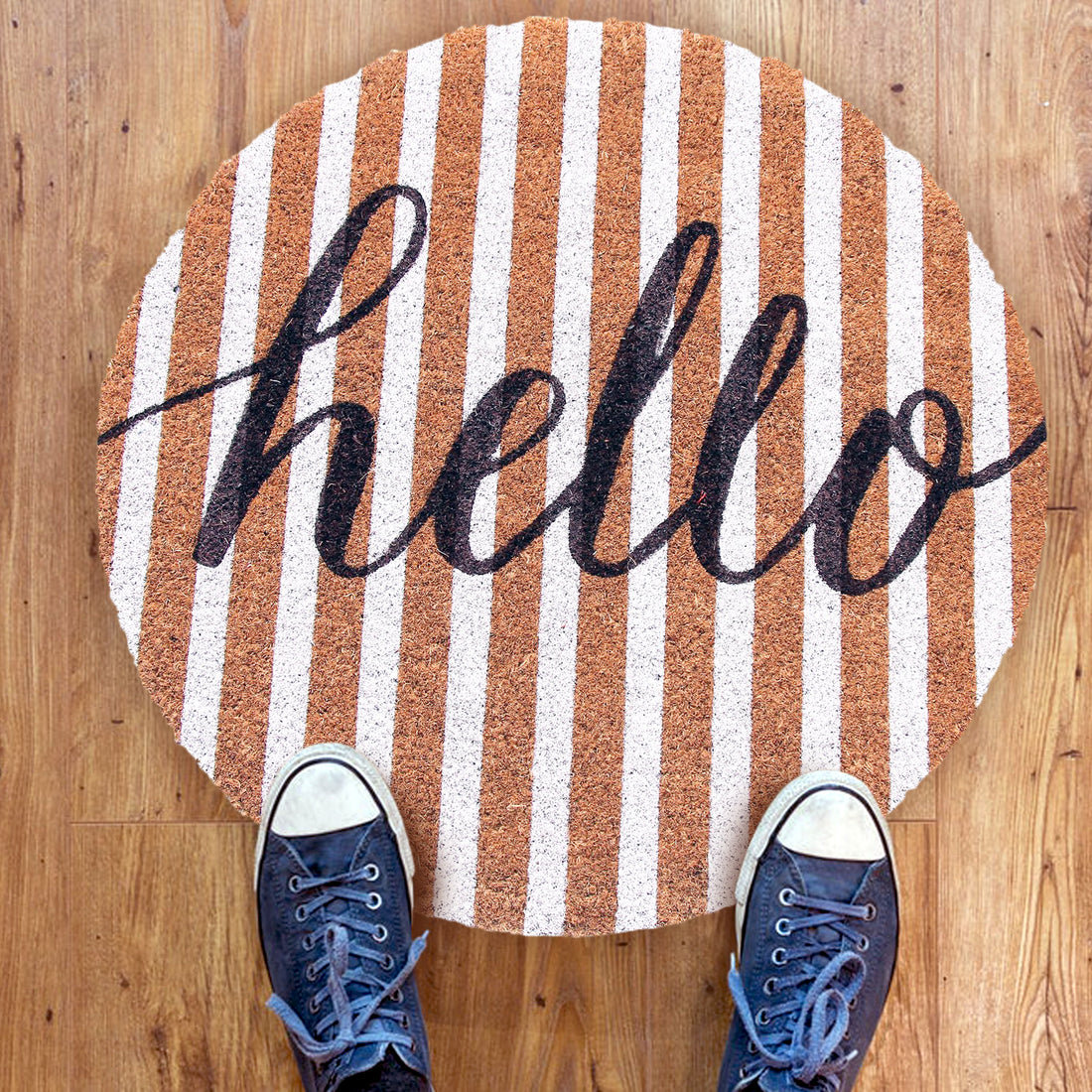 A round-shaped natural anti-slip coir floor mat with 'Hello' text in bold script, placed on a wooden floor, and a pair of sneakers positioned on top of the mat.