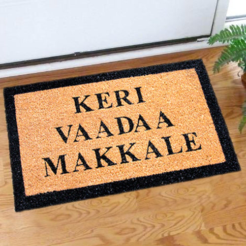 A rectangular natural coir doormat with black edging and the text 'KERI VAADA MAKKALE' printed in black. The mat is placed on a wooden floor, and there is a potted plant to the side.