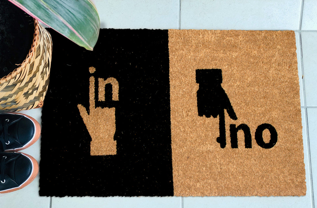 A rectangular natural coir entrance mat with a black and brown geometric pattern and text 'IN OUT', placed on a tiled floor next to a basket with a pair of shoes and a plant.
