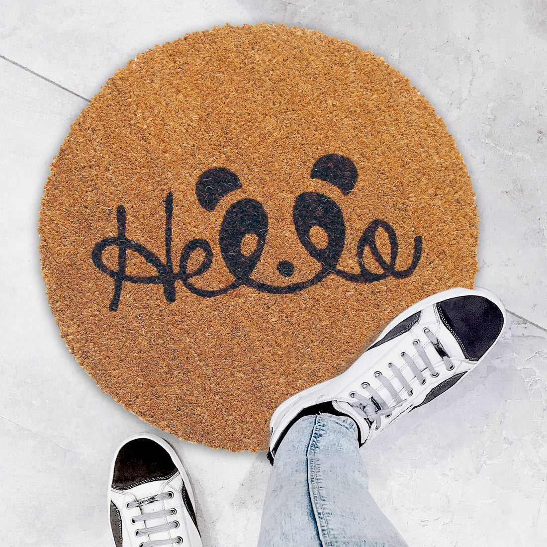 A round-shaped natural coir doormat with a black 'Hello' printed design, placed on the floor with a pair of sneakers at its top, and a person's leg and jeans visible to the side.