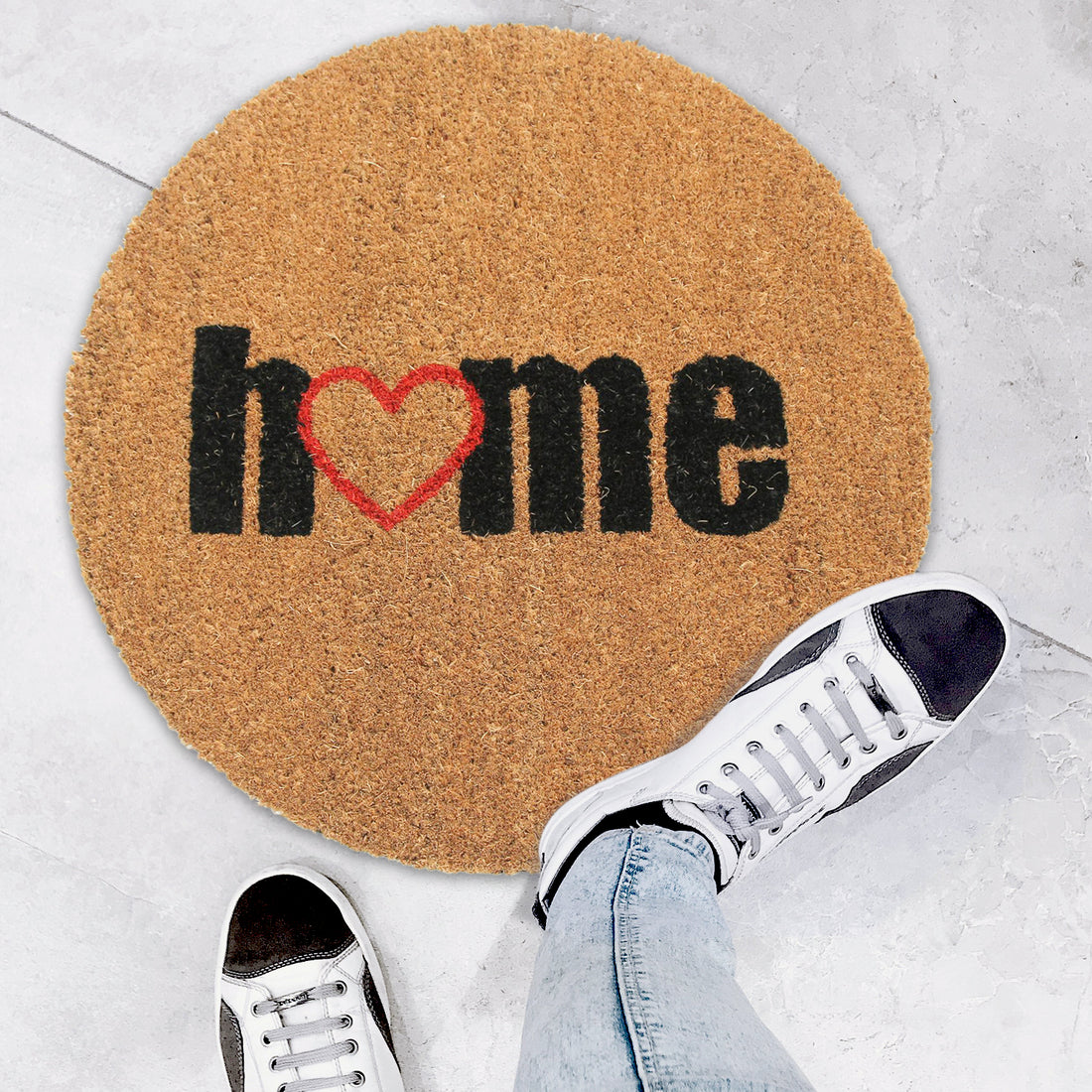 A round-shaped natural coir anti-slip doormat with the word 'Home' printed in black and a red heart symbol between the letters. The doormat is placed on the floor, and there are sneakers at the top of the image, indicating the scale of the mat.