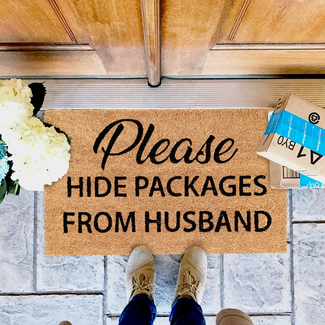 A rectangular natural coir floor mat with the text 'Please Hide Packages from Husband' printed on it, placed on the doorstep with a door in the background and a box hidden behind it.