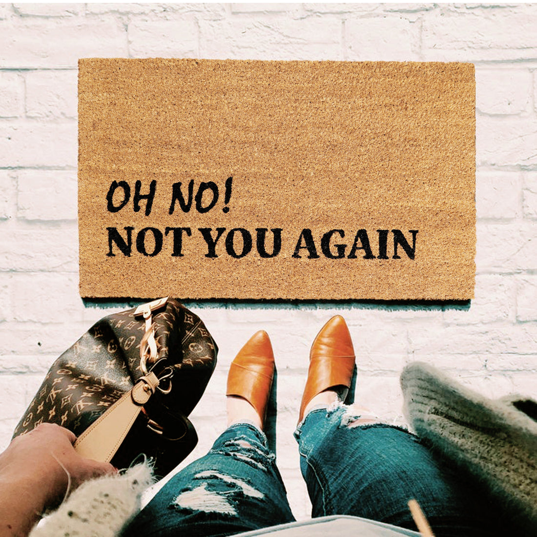 A natural coir door mat with a humorous text 'OH NO! NOT YOU AGAIN' printed on it, placed on a white brick floor, with a person's feet in shoes and jeans visible at the bottom of the image, indicating movement towards the mat.