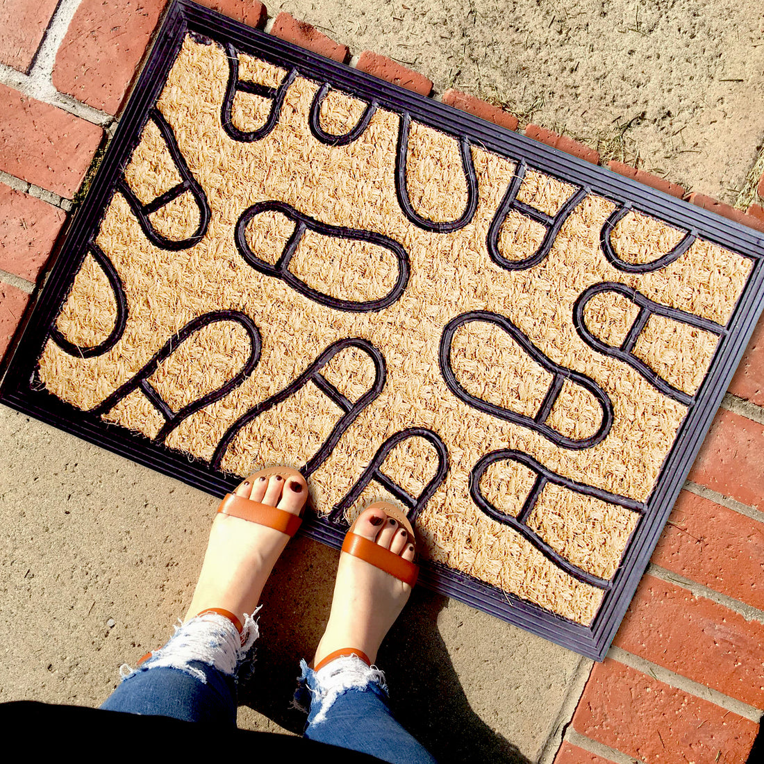 
                      
                        A black rubber door mat with a footprint design, moulded over a woven natural coir textile, with a person's feet placed on it, indicating the mat's use.
                      
                    