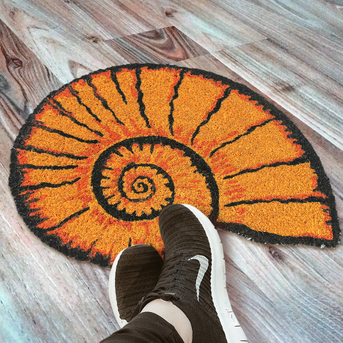 A round, orange anti-slip natural coir floor mat with a seashell shape, placed on a wooden floor, with a person's foot positioned on the mat wearing black sneakers.