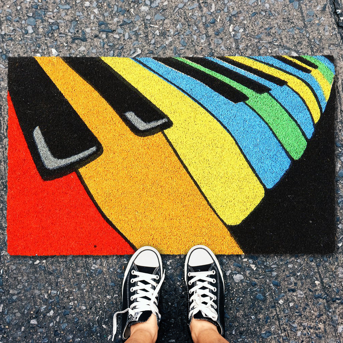 A rectangular coir door mat with a colourful printed design of a piano keyboard on it, placed on the ground with a pair of sneakers at the bottom of the image.