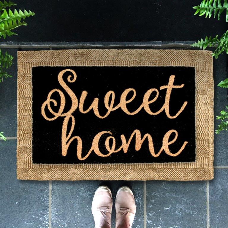 A black entrance doormat with the text 'Sweet Home' in a cursive font, placed on a doorstep with a person's feet in pink shoes over it.