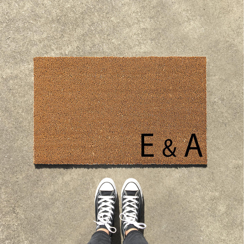 A rectangular personalised doormat with the letters 'E' and 'A' in the bottom right corner, in a brown colour, with a pair of sneakers on top of it.