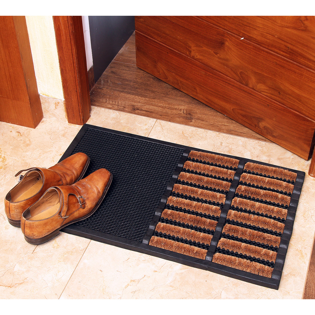 A black rubber wire brush door mat with cylindrical coir brushes, placed on the floor in front of a wooden door, with a pair of shoes resting on top of it.