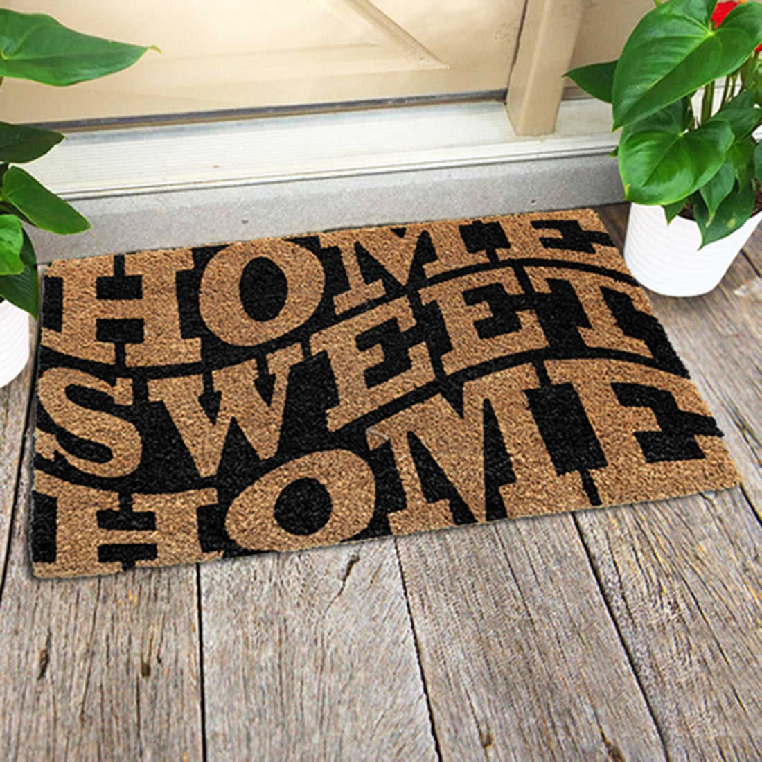 A rectangular coir doormat with the text 'HOME SWEET HOME' printed in black, placed on a wooden floor by a doorstep, with potted plants on either side.