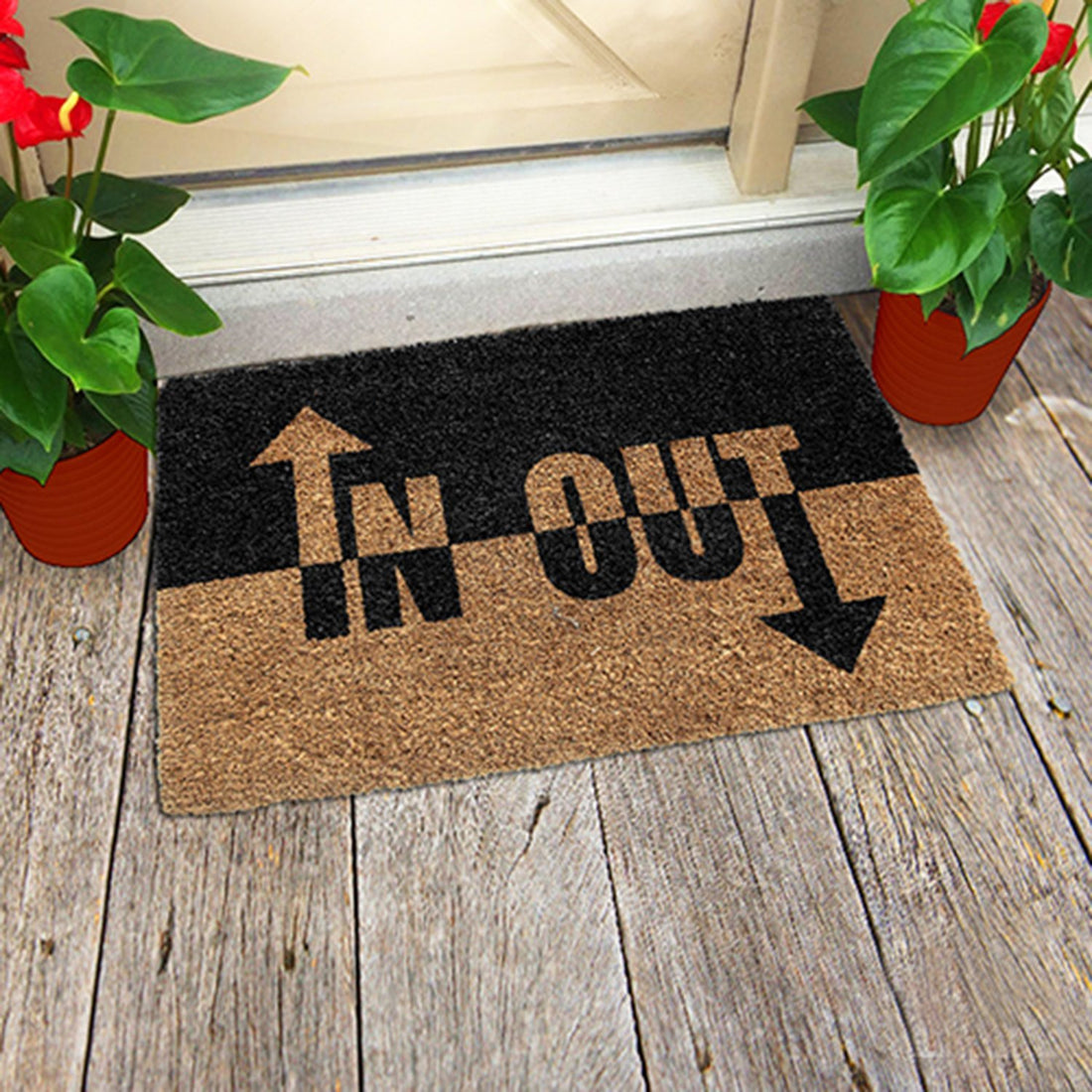 A rectangular coir doormat with a black and white 'IN OUT' printed design, placed on a wooden floor, with potted plants on either side.