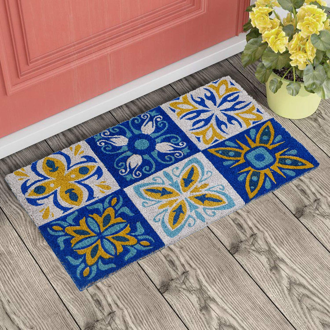 A rectangular multicoloured coir doormat with a geometric pattern, placed on a wooden floor with a flower pot beside it.
