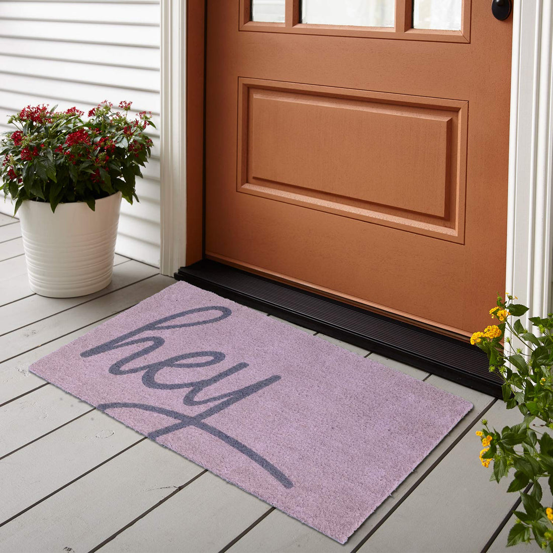 A rectangular natural coir door mat with the word 'Hey' printed in a different colour, placed on a wooden doorstep with a brown door and a black frame.