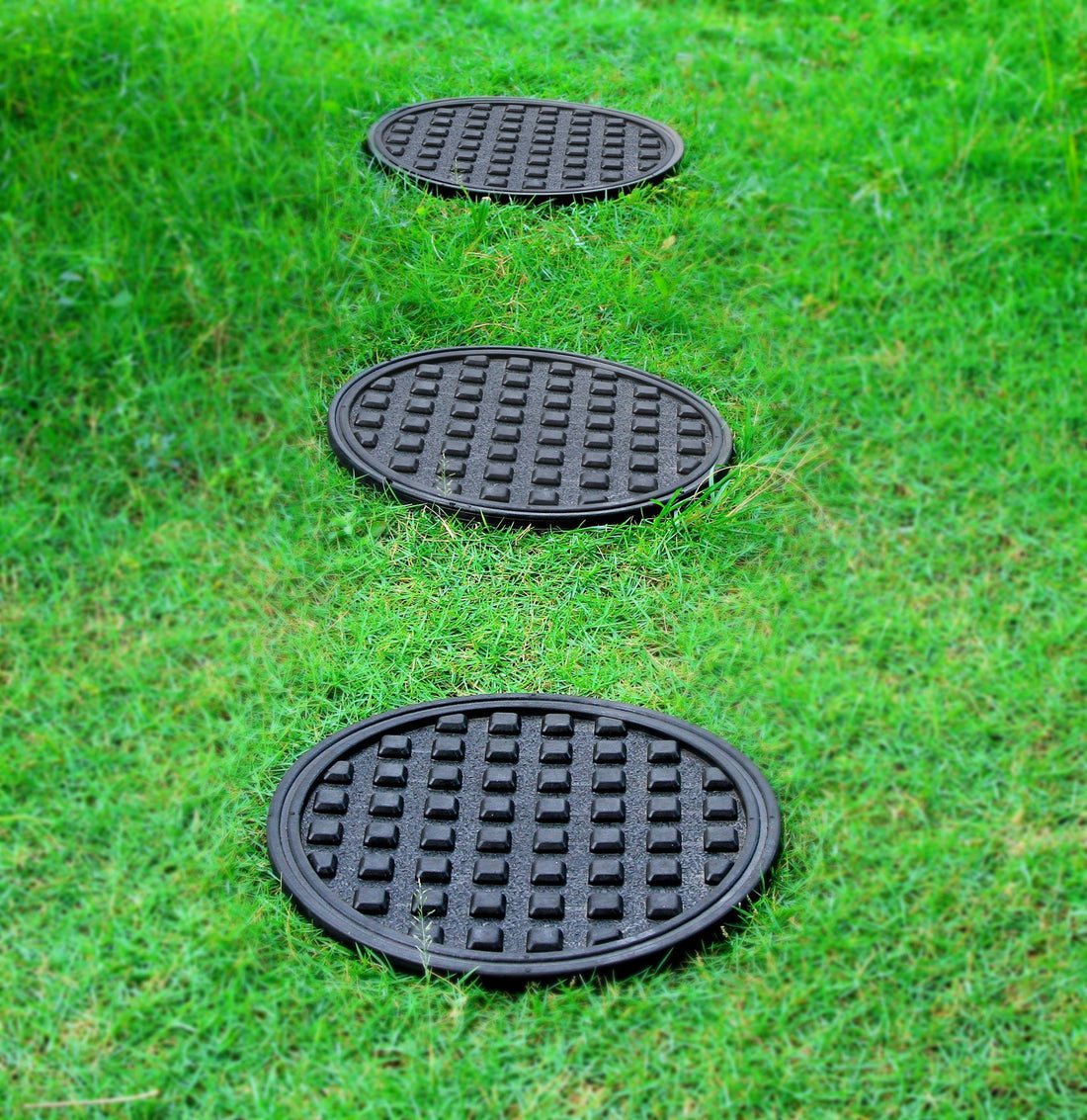 Three black round rubber garden tiles placed on grass, designed for use as a coaster on flower pots for indoor or outdoor plants.
