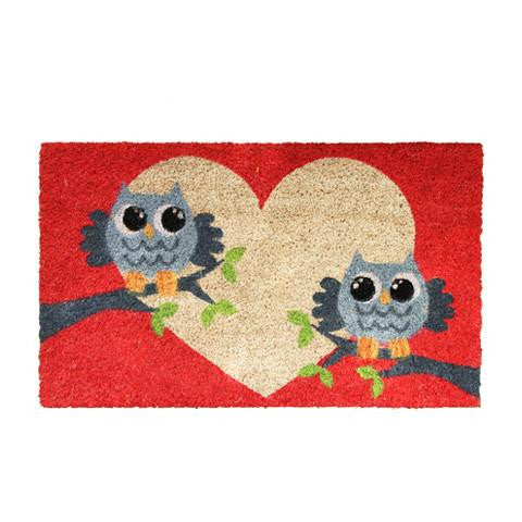 A rectangular coir door mat with a red background featuring two cartoon owls and a heart shape in the center.