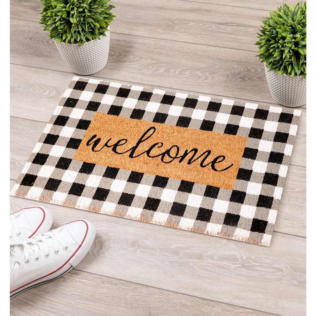 A natural coir doormat with a black and white checkered pattern and the word 'WELCOME' printed in black on a white background, placed on a wooden floor, accompanied by a pair of sneakers and two potted plants.