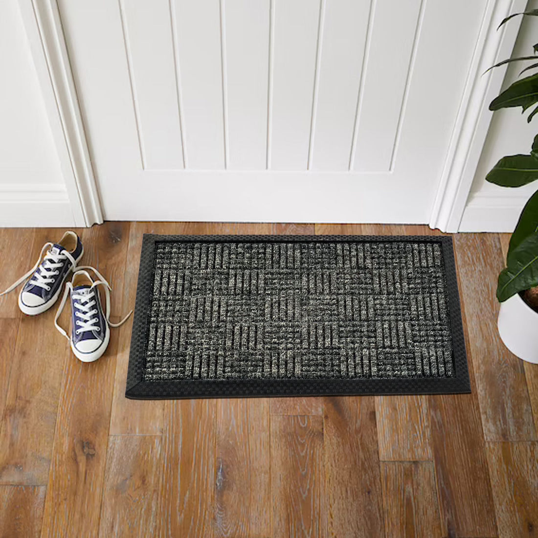 A charcoal grey, solid-patterned indoor rug is placed on a wooden floor, with a pair of sneakers at its top left corner and a potted plant to the side.