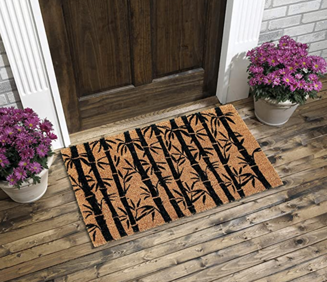 A rectangular natural coir doormat with a black and brown bamboo leaf design, placed on a wooden floor in front of a door, with potted plants on either side.