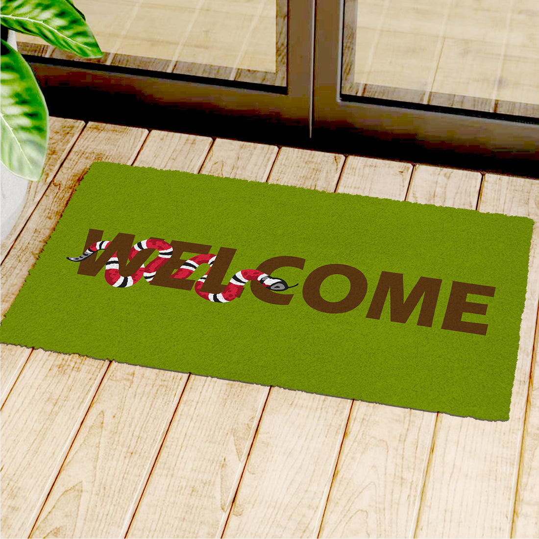 A green, natural coir entrance mat with a printed design featuring a snake and the word 'WELCOME' is placed on a wooden floor by a doorstep.