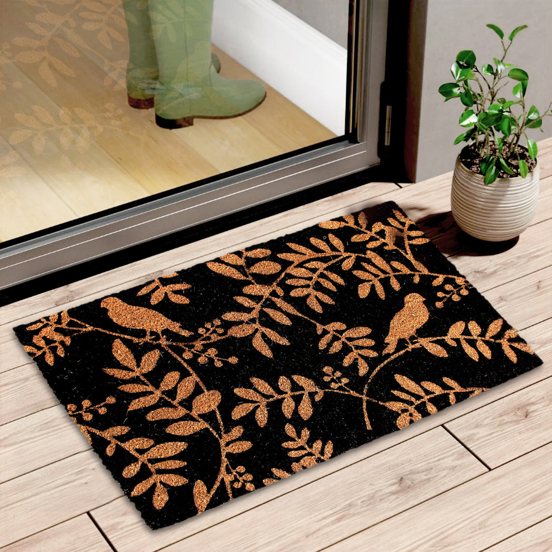 A rectangular black door mat with a leaf and bird design, placed on a wooden floor in front of a door, with a person's foot visible in a green bootied shoe, and a potted plant to the side.