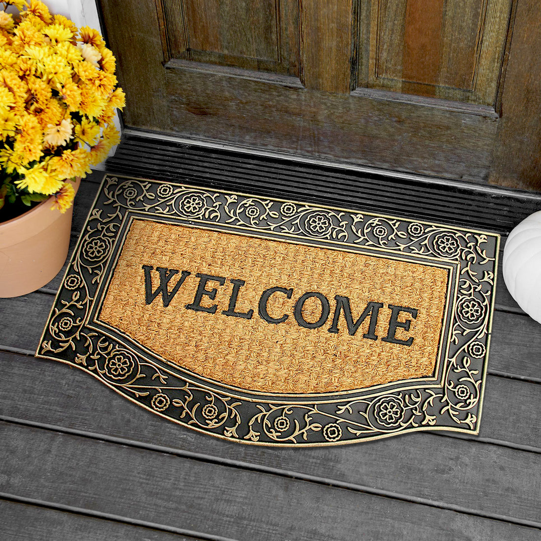 A natural coir doormat with a golden border, featuring an engraved design, placed on a wooden doorstep, with a potted plant and a wooden fence in the background.