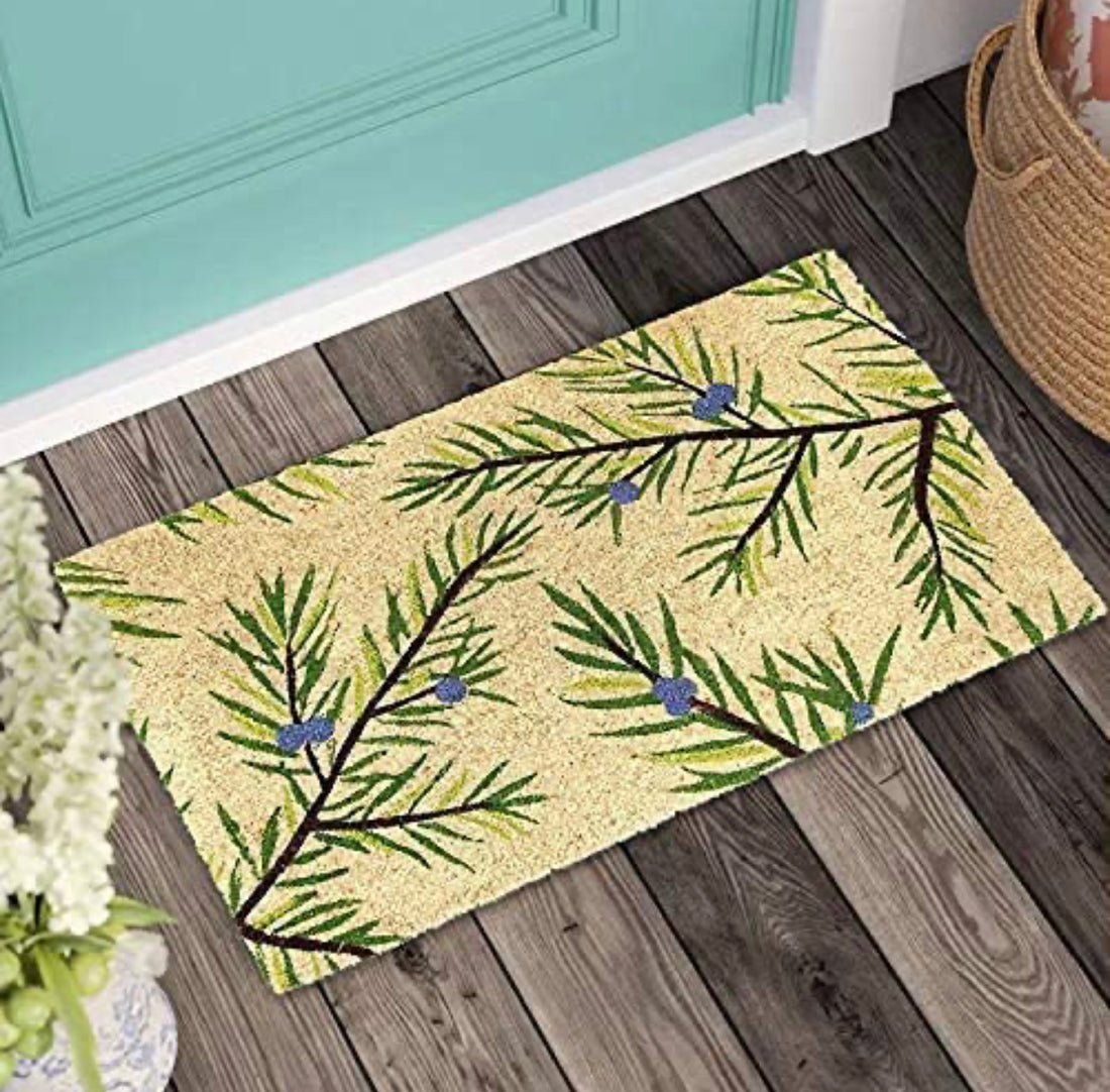 
                      
                        A rectangular beige and green coir doormat with a floral design, featuring a pattern of coconut trees and leaves, placed on a wooden floor with a wicker basket and a vase with flowers in the background.
                      
                    